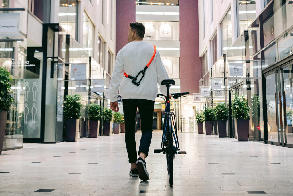 Radfahrer schiebt Fahrrad durch Passage mit Bügelschloss-Verlängerung tex-lock eyelet orange um die Schulter
