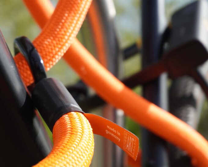 D-lock Bügelschloss sichert Sattelstange und Sattelstrebe mit Textilseil in Farbe acid orange und Webetikett "Made in Germany, ART*3" am Fahrradständer