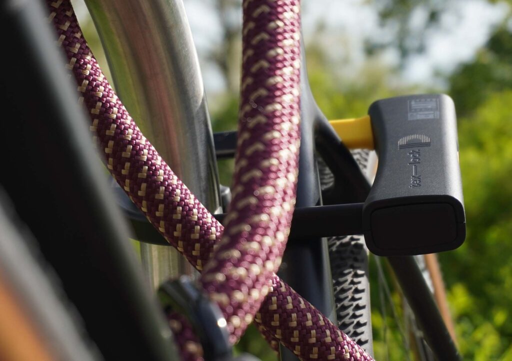 D-lock Bügelschloss sichert Sattelstange und Sattelstrebe mit Textilseil in Farbe chateau red am Fahrradständer