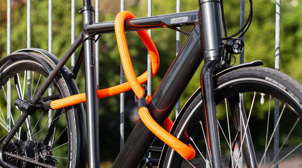 Flexible Bügelschloss-Verlängerung tex-lock eyelet L in orange sichert Rahmen und Räder mit Loop-Through-Prinzip und X-Lock an Geländer