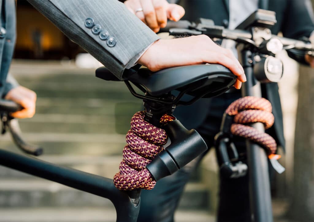 Model in grauem Sacko hält Fahrrad am Sattel, an dessen Stange das tex–lock orbit in chateau red flexibel gewickelt ist