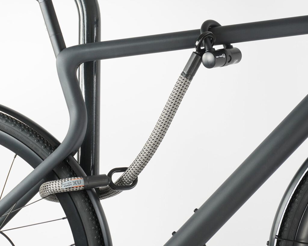 Hinterrad und Oberrohr vom Fahrrad mit Bügelschloss und Verlängerung aus Textil in grau-schwarz am Fahrradständer angeschlossen