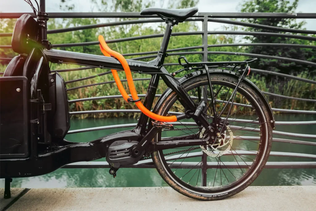 tex–lock eyelet in acid orange sichert schwarzes Lastenrad mit U-lock an Brückengeländer, Fluss im Hintergrund