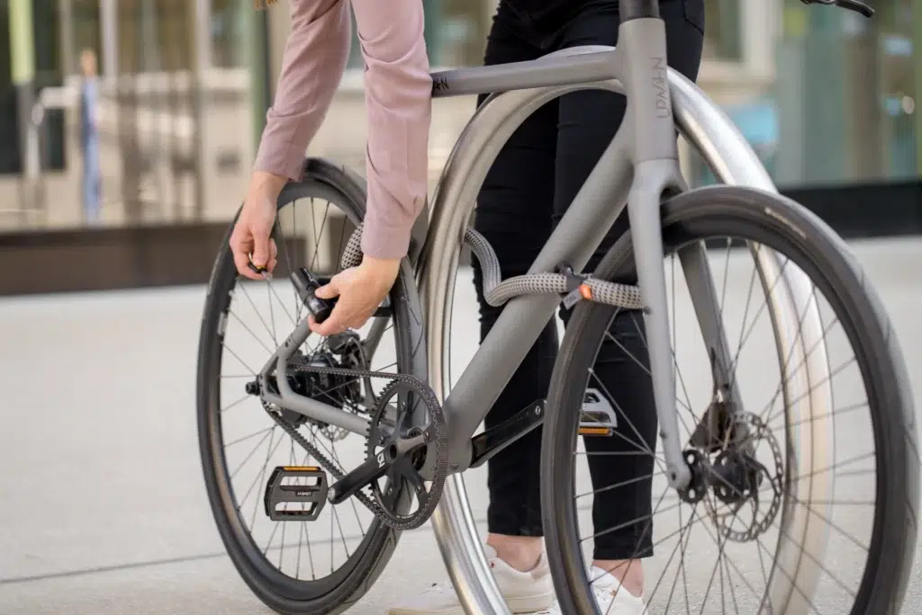 Frau in Anzughose und Hemd schließt Fahrrad mit tex–lock eyelet in electric grey als Leasing-Schloss an Fahrradstander ab.